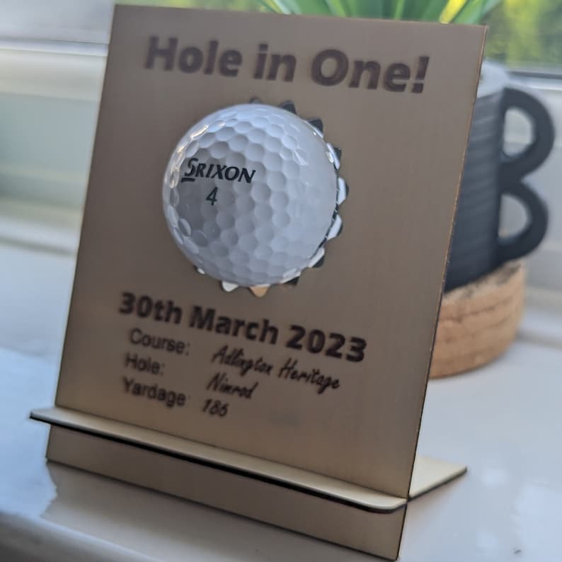 May include: A wooden plaque with a golf ball embedded in it. The plaque reads "Hole in One!" and includes the date "30th March 2023", the course "Addington Heritage", the hole "Nimrod", and the yardage "186".