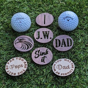 May include: Golf balls and wooden ball markers on green grass. The markers have various designs and text, including "I", "J.W.", "DAD", "Sink it", "Pops", and an eagle. The golf balls are branded Titleist and Callaway.