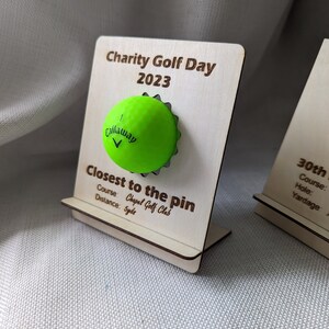 May include: A wooden plaque with the text "Charity Golf Day 2023" and "Closest to the pin" with a green golf ball attached. The plaque also includes the text "Course: Chapel Golf Club" and "Distance: 5yds".