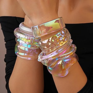 Peut inclure: Plusieurs bracelets en acrylique transparent et irisé. Les bracelets présentent divers motifs, lisses, texturés et en forme de bulles. La lumière se reflète sur les bracelets, créant un effet arc-en-ciel. Les bracelets sont empilés sur le poignet.