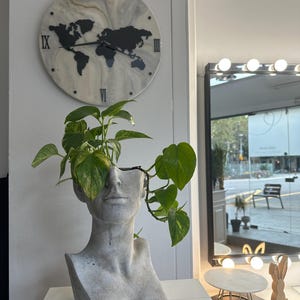 Puede incluir: Un macetero de busto de concreto gris con una planta verde que crece desde la parte superior de la cabeza. El busto está sentado sobre una superficie blanca. Un reloj con un diseño de mapa del mundo está en la pared detrás del macetero.