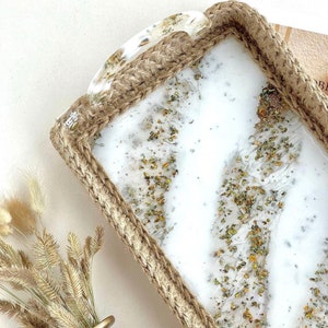 Puede incluir: Una bandeja rectangular blanca con un borde de cuerda de yute y un asa transparente. La bandeja tiene un fondo blanco con purpurina dorada incrustada en la resina.