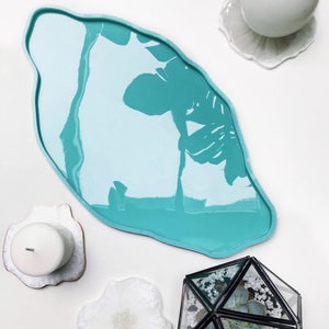 Puede incluir: Una bandeja de resina azul turquesa con una forma irregular y orgánica. La bandeja está sobre una superficie blanca con varios posavasos blancos y un pequeño terrario de vidrio con una planta suculenta.
