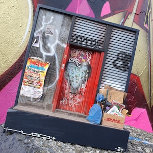 May include: A miniature model of a brick building with a red door and graffiti on the walls. The building is sitting on a black base with the word "HUGO" written on it. There are cardboard boxes and a blue bag in front of the building.