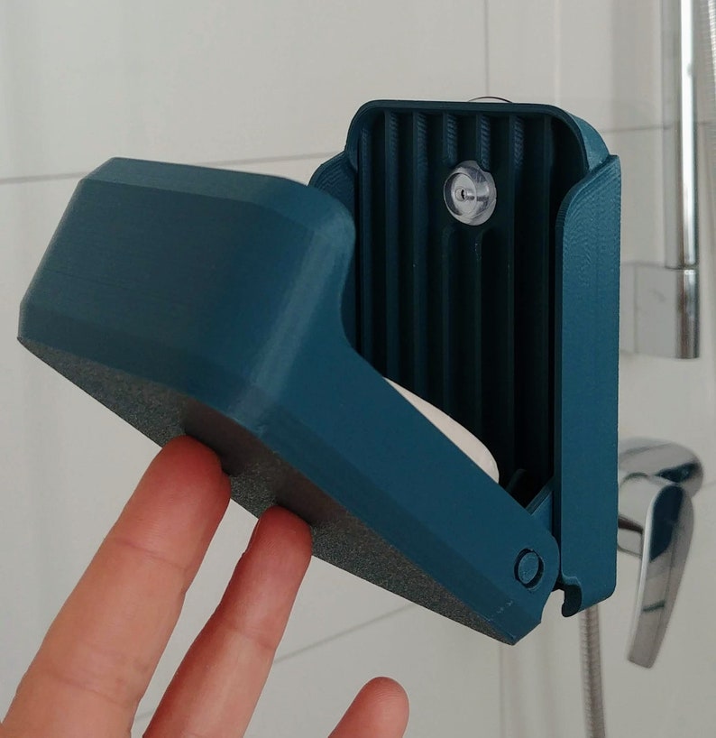 One hand closing a 3d printed blue soap box under the shower, containing solid shampoo. It hangs from a suction cup. Wall mounted