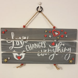 May include: A grey wooden sign with the text "Love Changes Everything" in white paint. The sign is decorated with red hearts and gold stars. The sign is hanging from a rope with red beads.