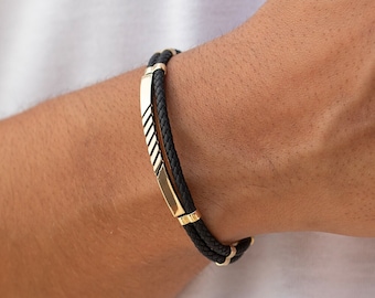 May include: A black leather bracelet with a gold metal accent. The bracelet has a braided design and is worn on a person's wrist.