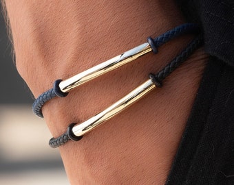 May include: Two bracelets with braided leather bands and gold-coloured cylindrical bars. One bracelet is navy blue, the other is black. The bracelets are worn on a person's wrist, with the person's arm and clothing visible.