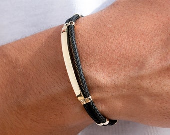 May include: A black leather bracelet with a gold bar and gold accents. The bracelet is braided and has a simple design.