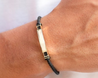 May include: A black leather bracelet with a gold bar and black accents. The bar has a white inlay.