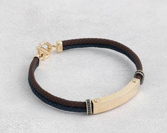May include: A brown and blue braided leather bracelet with a gold bar and black accents. The bar has a textured surface and is secured with screws.