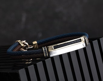 May include: A brown, blue and black leather bracelet with a silver and gold bar accent. The bar has black gemstones on either side.