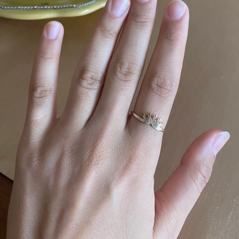 K&ouml;nnte beinhalten: Ein goldfarbener Ring mit einem Sonnenstrahlen-Design. Der Ring wird an einem Finger getragen und zeigt die feinen Details der Sonnenstrahlen. Das Design des Rings ist minimalistisch und elegant, geeignet f&uuml;r den t&auml;glichen Gebrauch.