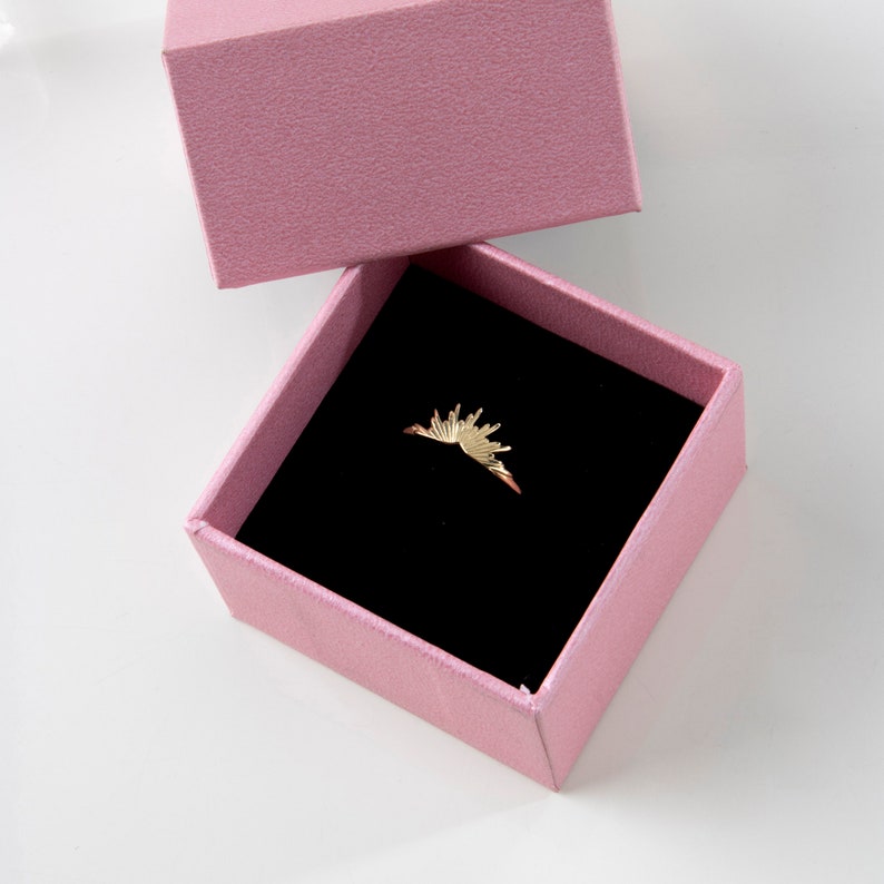 K&ouml;nnte beinhalten: Ein goldfarbener Ring mit Sonnenstrahlen-Design, pr&auml;sentiert in einer rosa Schmuckschatulle mit schwarzem Innenraum. Die Schatulle ist ge&ouml;ffnet und zeigt die filigranen Details des Rings. Der Ring ist ein zartes Schmuckst&uuml;ck.