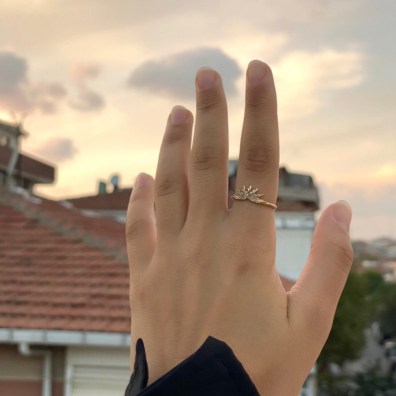 K&ouml;nnte beinhalten: Ein goldfarbener Ring mit einem Sonnenstrahlen-Design. Der Ring wird an einem Finger getragen, wobei das Sonnenstrahlen-Detail nach oben zeigt. Der Hintergrund zeigt einen bew&ouml;lkten Himmel und D&auml;cher.