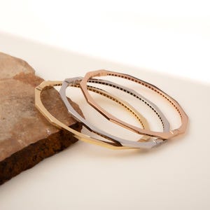 Könnte beinhalten: Vier geometrische Armreifen in Gold-, Silber- und Roségoldtönen. Die Armbänder haben ein facettiertes Design und sind auf einem neutralen Hintergrund angeordnet. Die Armreifen sind ein stilvolles Accessoire.