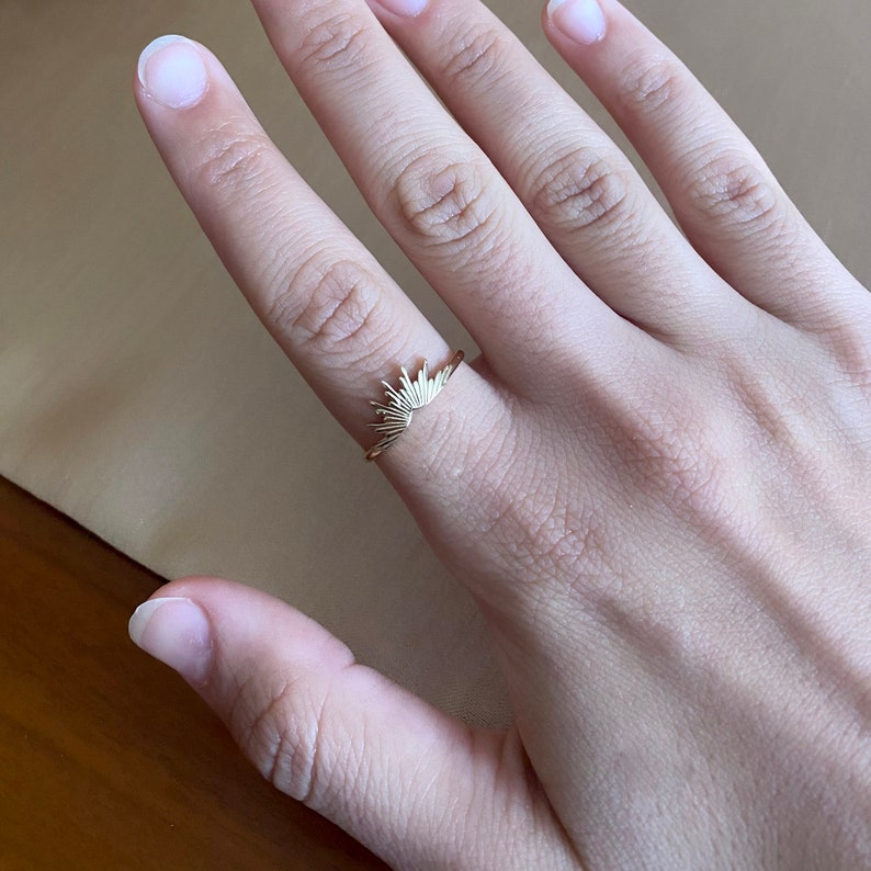 K&ouml;nnte beinhalten: Ein goldfarbener Ring mit einem Sonnenstrahlen-Design. Der Ring befindet sich an einem Finger und zeigt die feinen Details des sonnenartigen Musters. Das Design des Rings ist minimalistisch und elegant.