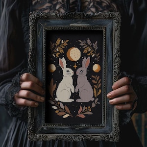 以下が含まれることがあります： 黒い背景に、白とグレーの2匹のウサギが手をつないでいるクロスステッチのアートワーク。デザインには、金、茶色、クリーム色の月、星、葉が含まれており、暗く装飾的なフレームに収められています。