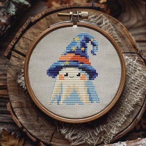 May include: Cross-stitch embroidery of a smiling ghost wearing a blue and orange wizard hat with yellow stars. The embroidery is set in a wooden hoop and placed on a wooden slice with fall foliage.