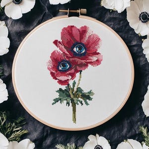 以下が含まれることがあります： 青い目をした赤いアネモネ2つがデザインされたクロスステッチ刺繍のフープ。アネモネは白い花に囲まれています。