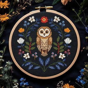 May include: A cross-stitch embroidery hoop featuring a detailed owl design. The owl is centered on a black fabric, surrounded by a floral wreath with white, orange, and blue accents. The wooden hoop is approximately 8 inches in diameter.