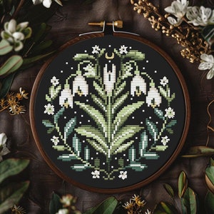 May include: A cross-stitch embroidery hoop with a floral design. The design features white flowers, green leaves, and a crescent moon on a black background. The embroidery is set in a wooden hoop, surrounded by greenery.