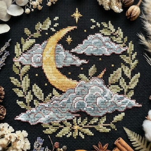 Pode incluir: Bordado em ponto cruz sobre tecido preto com uma lua crescente, nuvens, estrelas e folhagem. A lua é dourada, as nuvens são cinza-azuladas e a folhagem é verde. O desenho é rodeado por flores secas e especiarias.