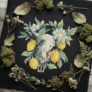 May include: Cross-stitch design on black fabric featuring a skull decorated with lemons, white flowers, and green leaves. The design is surrounded by dried leaves, moss, and sprigs. The colour palette includes yellow, white, green, and black.