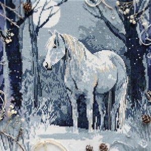May include: Cross-stitch artwork of a white horse in a snowy forest under a full moon. The piece uses blue and white threads, with detailed stitching creating a textured effect. Pine cones and twigs frame the scene.
