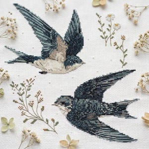 Könnte beinhalten: Eine Kreuzstichstickerei auf weißem Stoff mit zwei fliegenden Schwalben in Dunkelblau-, Grau- und Cremetönen. Das Design enthält zarte, gestickte Zweige mit kleinen Blumen und Grünpflanzen, die zwischen den Vögeln angeordnet sind.