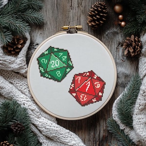 Puede incluir: Primer plano de un aro de bordado blanco con dos dados bordados en él. Los dados son verde y rojo, con números blancos. El aro está sobre una superficie de madera con ramas de pino y piñas.