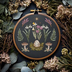 Puede incluir: Un bastidor de bordado de punto de cruz con un dise&ntilde;o tem&aacute;tico de la naturaleza. El dise&ntilde;o incluye flores blancas y rosas, &aacute;rboles verdes, setas marrones y un huevo moteado en un nido, todo sobre un fondo gris oscuro. El bastidor de madera est&aacute; rodeado de plantas secas.