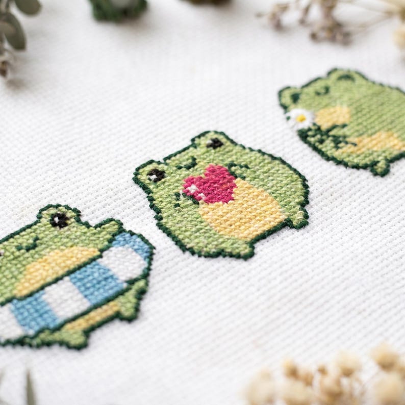 May include: A cross-stitch embroidery on white fabric featuring three small green frogs. One frog holds a blue and white striped object, the center frog holds a pink heart, and the third frog holds a small white flower with a yellow center.