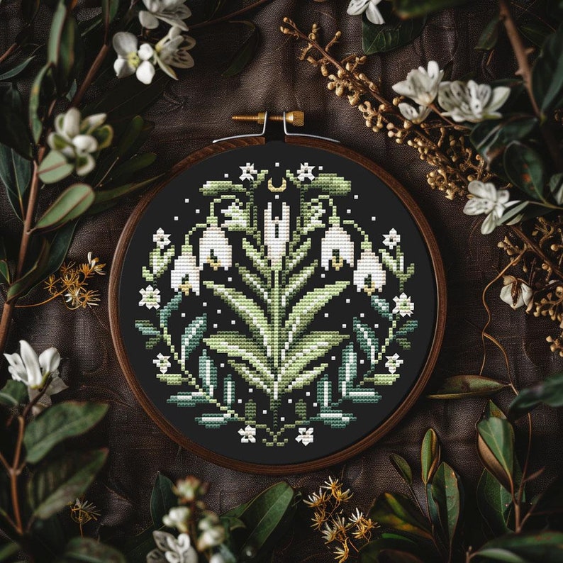 May include: A cross-stitch embroidery hoop with a floral design. The design features white flowers, green leaves, and a crescent moon on a black background. The embroidery is set in a wooden hoop, surrounded by greenery.