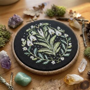 May include: A circular cross-stitch embroidery on black fabric, depicting white flowers and green foliage, set in a wooden hoop. The image is surrounded by crystals, moss, and pine cones. The embroidery hoop is approximately 15 cm in diameter.