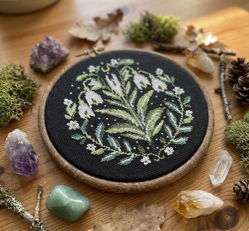 May include: A circular cross-stitch embroidery on black fabric, depicting white flowers and green foliage, set in a wooden hoop. The image is surrounded by crystals, moss, and pine cones. The embroidery hoop is approximately 15 cm in diameter.