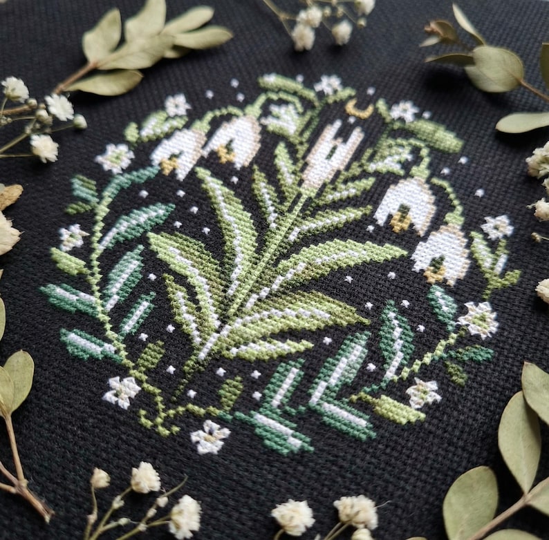 May include: A cross-stitch design of a floral wreath with white bell-shaped flowers, green leaves, and small white blossoms on a black background. The design is surrounded by dried flowers and leaves.