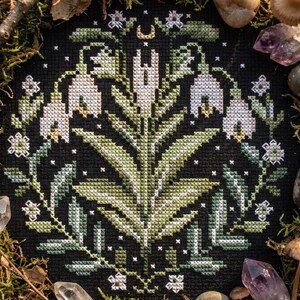 May include: A cross-stitch floral design with white and green flowers, leaves, and a crescent moon on a black backdrop. The embroidery is encircled by moss, crystals, and stones, creating a natural, earthy aesthetic. The design is a circular wreath.