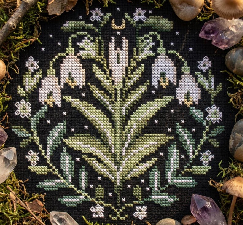 May include: A cross-stitch floral design with white and green flowers, leaves, and a crescent moon on a black backdrop. The embroidery is encircled by moss, crystals, and stones, creating a natural, earthy aesthetic. The design is a circular wreath.
