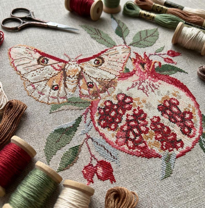 May include: A cross-stitch design depicting a moth and a pomegranate. The moth has tan and brown wings with red details. The pomegranate is red with green leaves. Spools of thread and scissors are visible in the background.