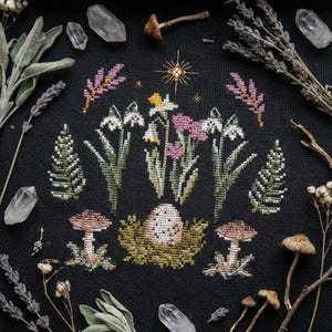 Puede incluir: Un dise&ntilde;o de punto de cruz sobre tela negra con un arreglo floral central con flores blancas, rosas y amarillas. El dise&ntilde;o incluye setas, un huevo en un nido y ramitas de lavanda y salvia. Cristales de cuarzo transparente y setas secas rodean el dise&ntilde;o.