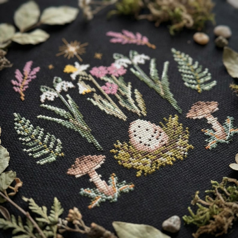 Puede incluir: Bordado de punto de cruz sobre tela negra con una escena de jard&iacute;n primaveral. El dise&ntilde;o incluye flores blancas y rosas, helechos verdes, setas y un huevo moteado en un nido. La paleta de colores es suave, con verdes, rosas y marrones apagados.
