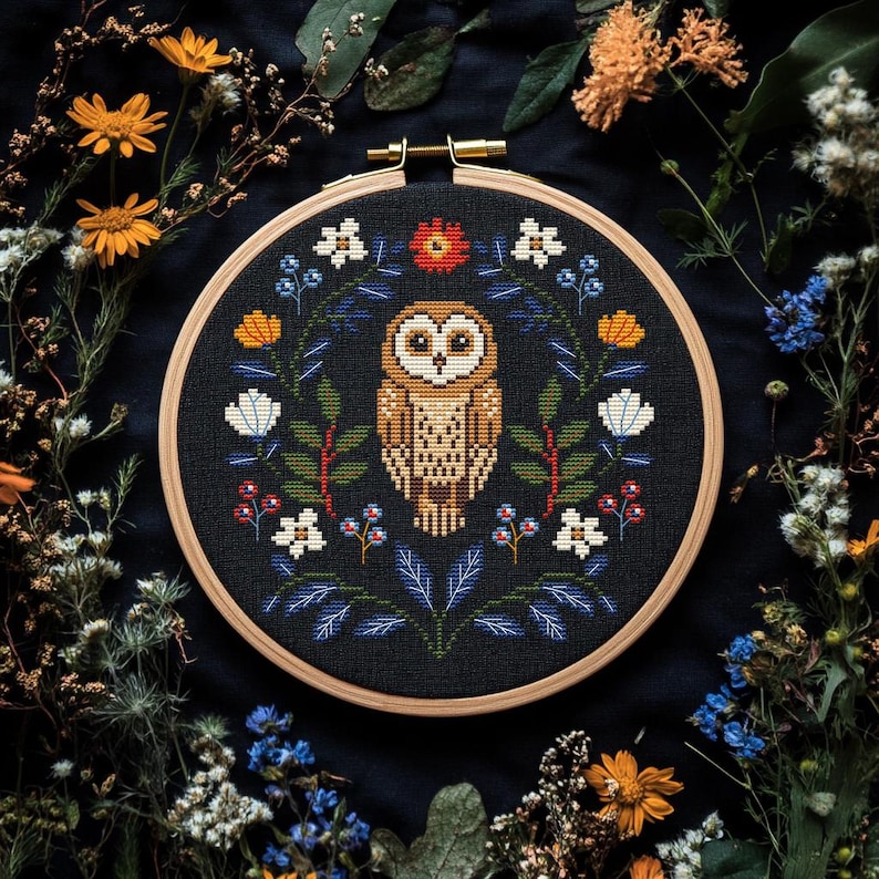 May include: A cross-stitch embroidery hoop featuring a detailed owl design. The owl is centered on a black fabric, surrounded by a floral wreath with white, orange, and blue accents. The wooden hoop is approximately 8 inches in diameter.