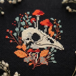 May include: A cross-stitch embroidery on black fabric featuring a white bird skull surrounded by orange and red mushrooms, small orange berries, and light blue foliage. The design is centred and composed of intricate, colourful thread stitches.
