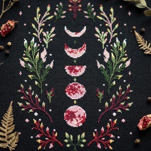 Op de afbeelding: Kruissteekontwerp met de maanfasen in roze en rood, omgeven door bloem- en botanische elementen in groen, roze en rood op een zwarte achtergrond. Het ontwerp bevat halve manen en cirkelvormen.