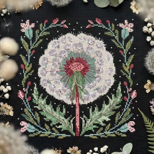 May include: Cross-stitch embroidery of a dandelion seed head, centered on a black background. The dandelion is white with a pink and teal center, surrounded by green leaves and pink flowers. The design is framed by a floral border.
