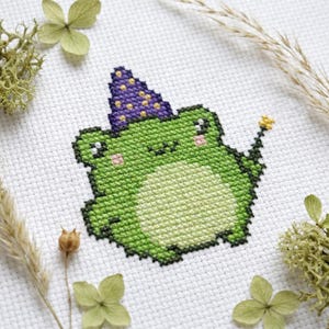 Puede incluir: Diseño de punto de cruz de una rana verde con un sombrero de mago morado con estrellas amarillas. La rana sostiene una varita de estrella amarilla. El diseño está sobre tela blanca, rodeado de flores y hierbas secas.