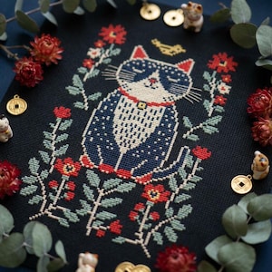 Könnte beinhalten: Kreuzstich-Stickerei einer Katze mit blau-weiß gemustertem Fell, rotem Kragen und floralem Rand auf schwarzem Hintergrund. Die Katze ist von roten Blumen, grünen Blättern und kleinen Glückskatzenfiguren umgeben. Goldmünzen und Zweige mit Grünzeug vervollständigen das Design.