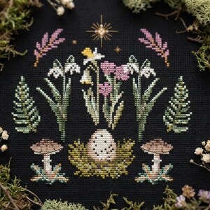 Puede incluir: Dise&ntilde;o de punto de cruz con tem&aacute;tica floral y de bosque. El dise&ntilde;o incluye flores blancas y rosas, setas y un huevo en un nido, todo sobre un fondo negro. El dise&ntilde;o est&aacute; enmarcado por musgo y flores secas.