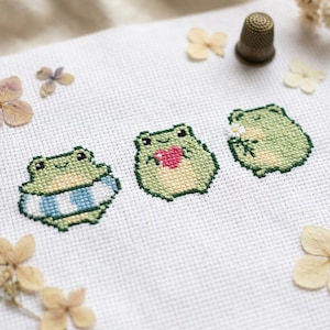 May include: A white Aida fabric featuring three cross-stitched green frogs. The first frog wears a blue and white striped inner tube, the second holds a pink heart, and the third holds a small white flower. A metal thimble sits nearby.